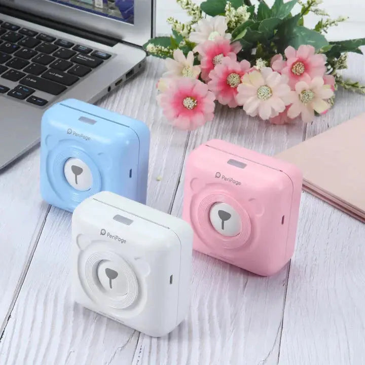 Pink blue and white compact portable mini printers on white wooden table near laptop and flowers